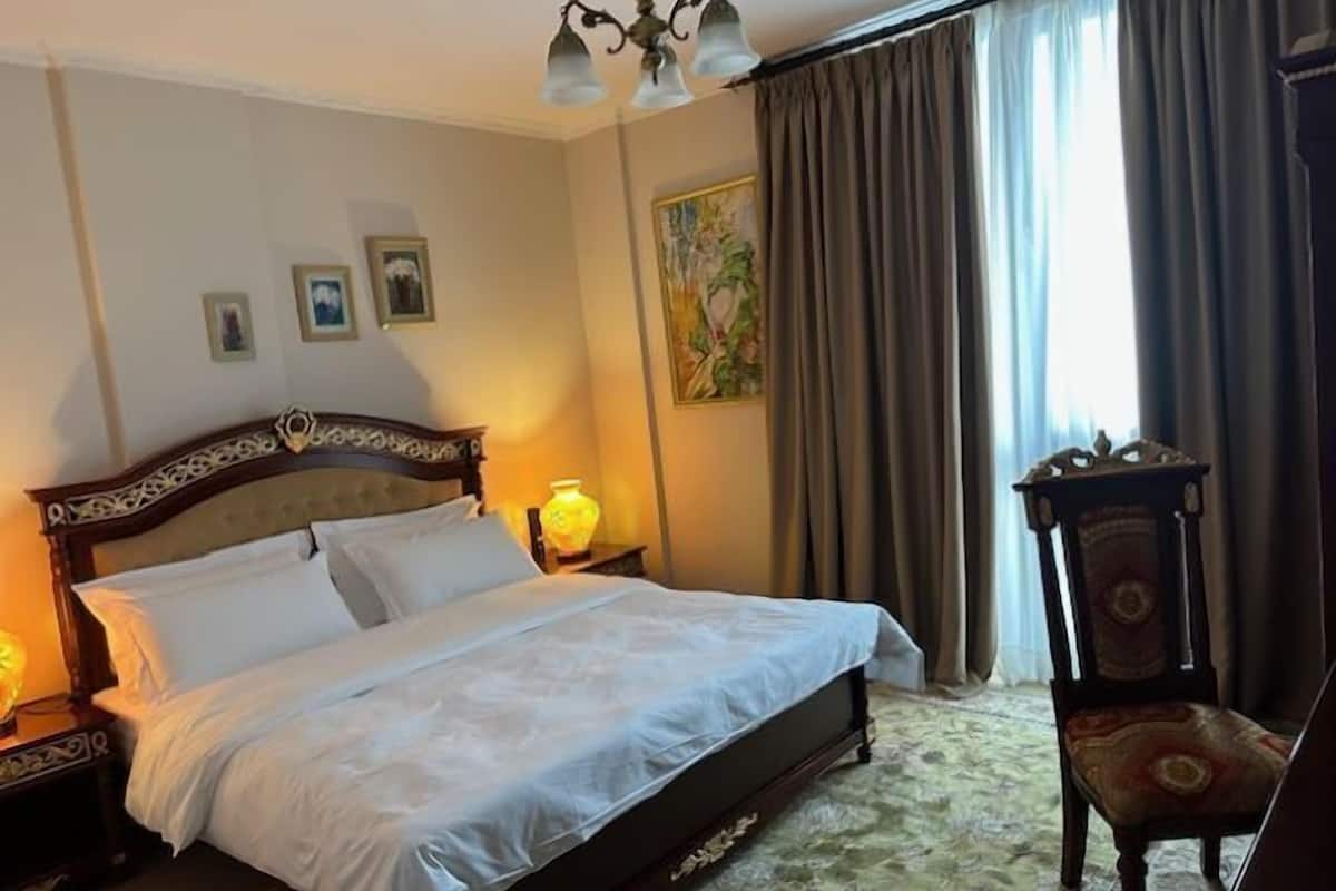 Een opgemaakt bed met witte lakens, een houten hoofdbord en twee bedlampjes. Er staat een stoel met een gestreept zitkussen en een decoratietafeltje naast het bed.
