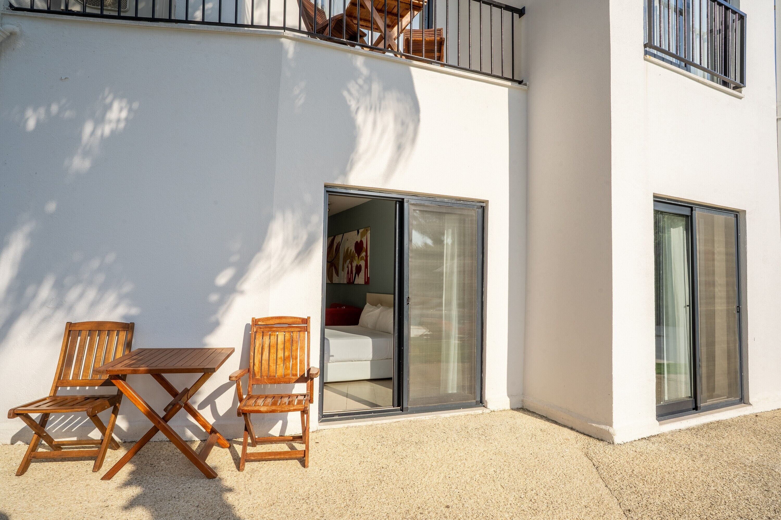 Um edifício moderno com varanda, mesa e cadeiras de madeira, e porta de correr de vidro que dá para um quarto com cama e um quadro.