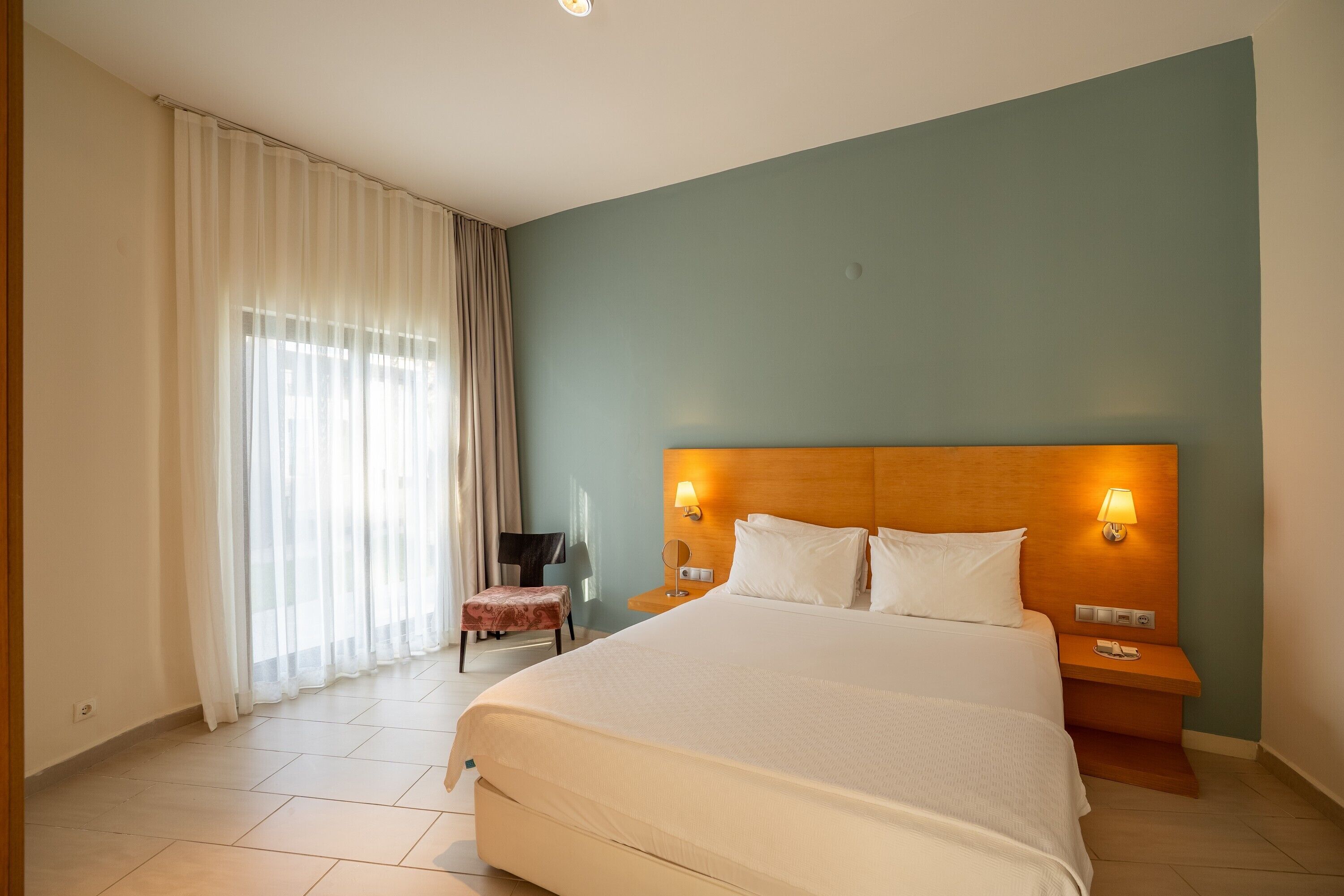 A hotel room with a large bed, two bedside lamps, a nightstand, a chair, and a window with curtains.