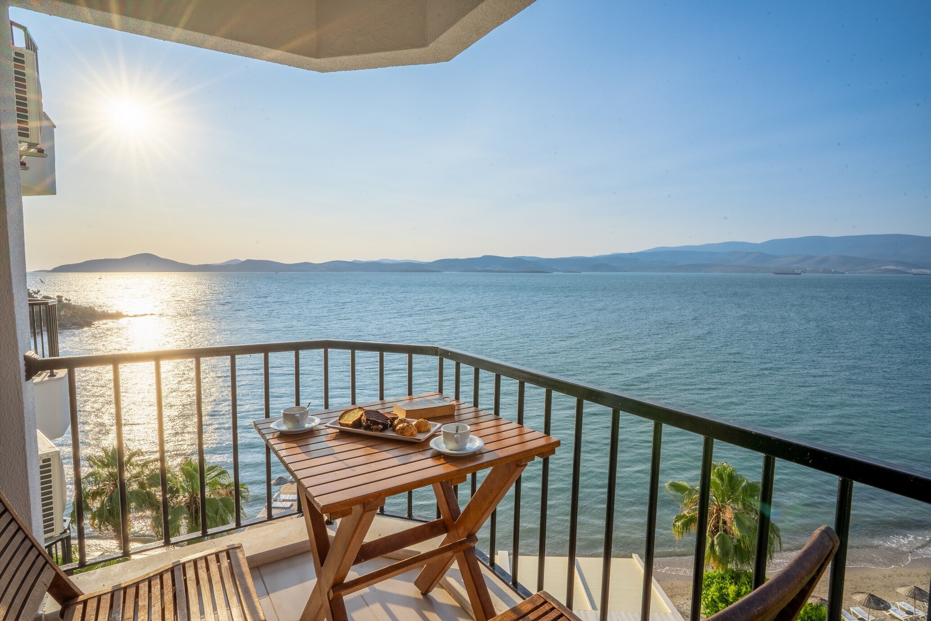 Uma varanda com mesa e cadeiras de madeira, com vista para um mar calmo e montanhas ao longe.