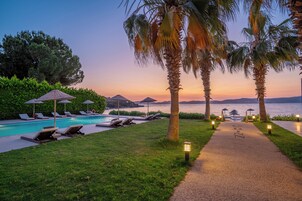 Un área junto a la piscina con sillones de descanso y sombrillas, un camino que lleva a la playa y vistas al atardecer.
