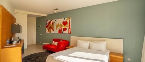 A hotel room with a large bed, a red armchair, a desk with a computer, and a painting on the wall.