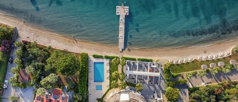 An aerial view of a coastal resort with a pier extending into the sea, a swimming pool, and a building with multiple balconies.