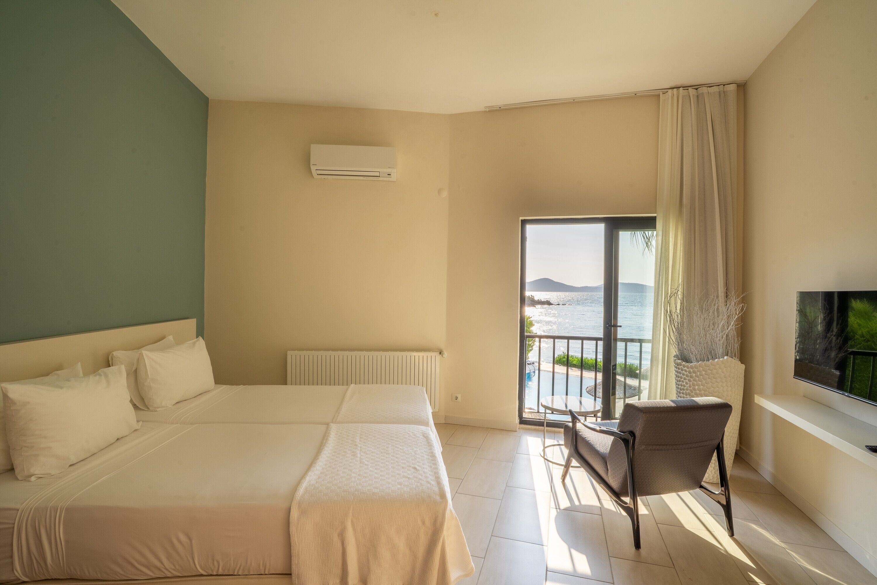 A hotel room with a large bed, a chair, a TV, and a balcony with a view of the sea.