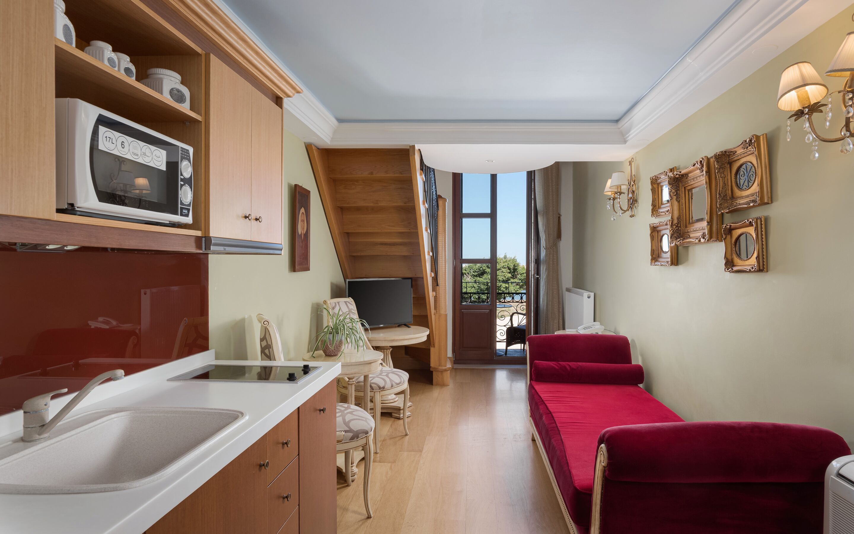 Cucina compatta con forno a microonde, lavello e pensili in legno. È presente un piccolo angolo ristoro con tavolo e sedie, e un divano rosso. La stanza dispone di un balcone con vista sul verde.