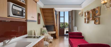 A compact kitchen with a microwave, sink, and wooden cabinets. There is a small dining area with a table and chairs, and a red sofa. The room has a balcony with a view of greenery.