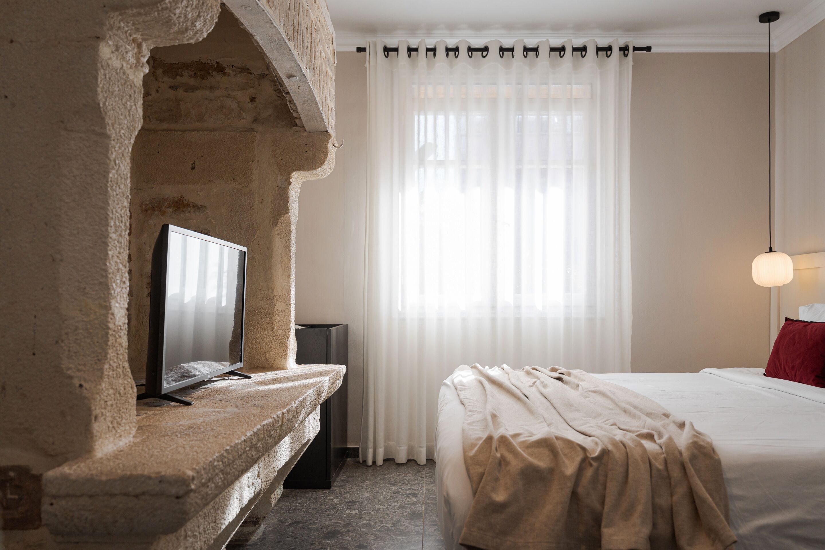 Ett hotellrum med en vägg som har en dekorativ stenbeklädnad, en platt-TV, en säng med en röd kudde och ett stort fönster med tunna, genomskinliga gardiner.