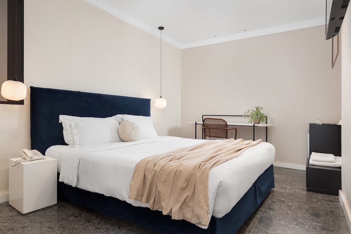 A modern hotel room with a large bed, bedside table, and a desk with a chair.
