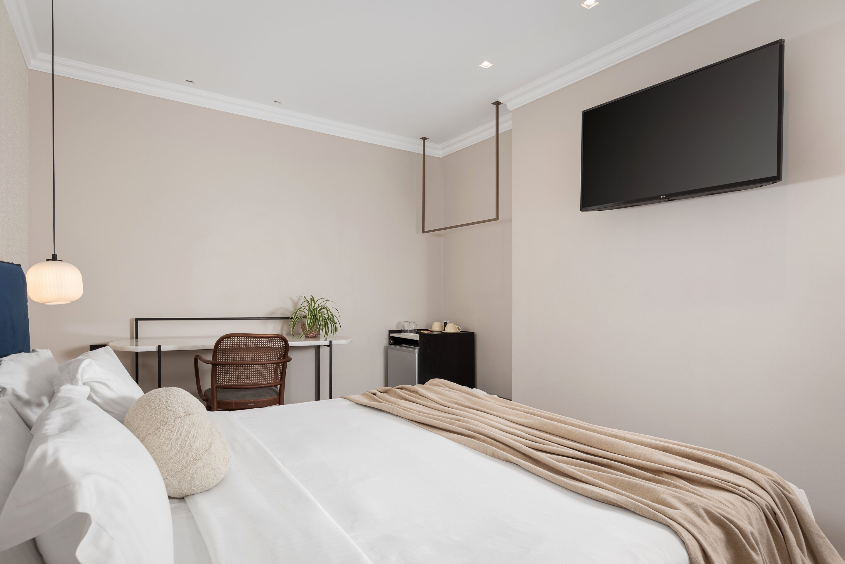 A hotel room with a bed, a desk, a chair, a TV mounted on the wall, and a hanging light fixture.
