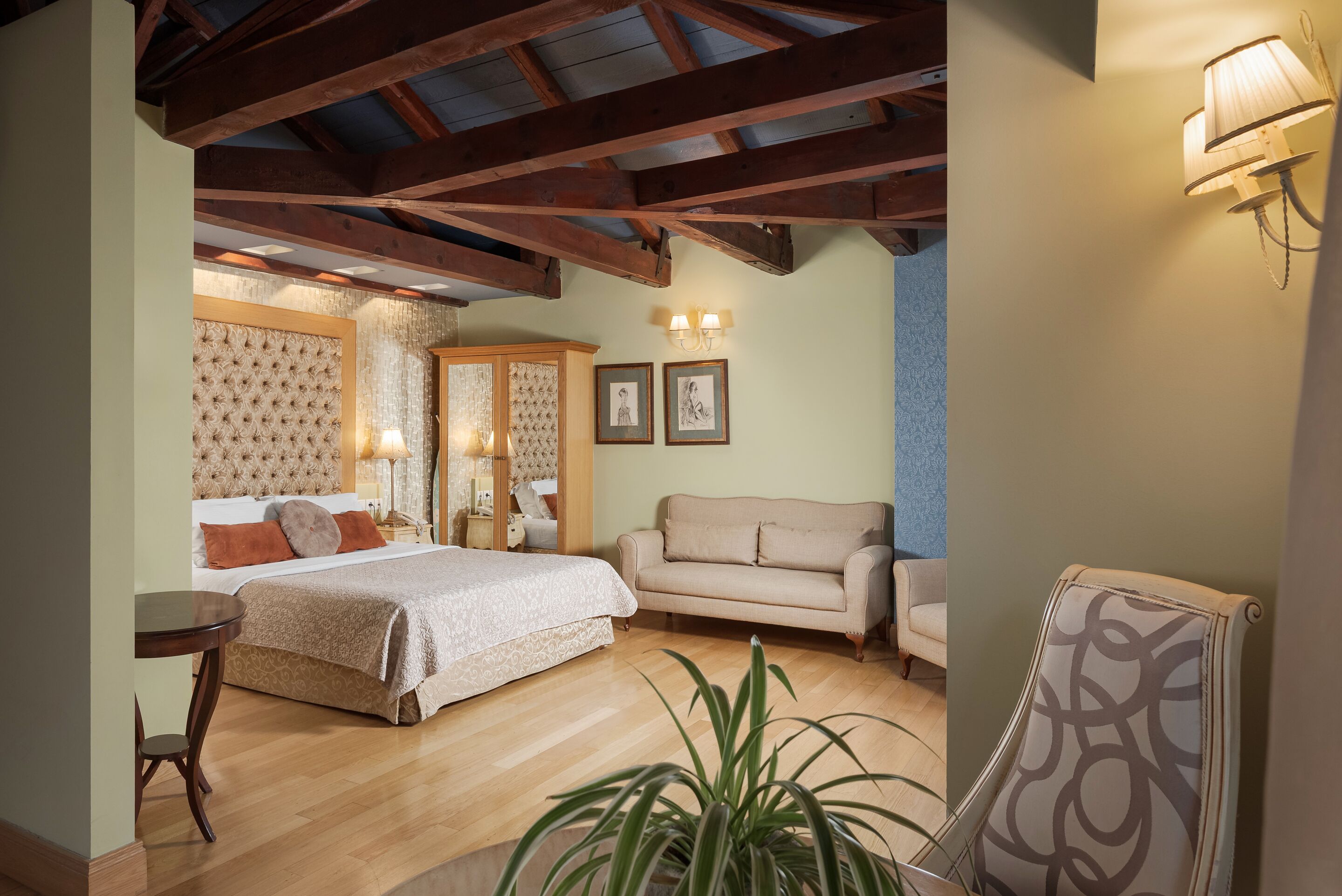 A bedroom with a large bed, a sofa, a small table, and a chair. There are two wall-mounted lamps and a potted plant.