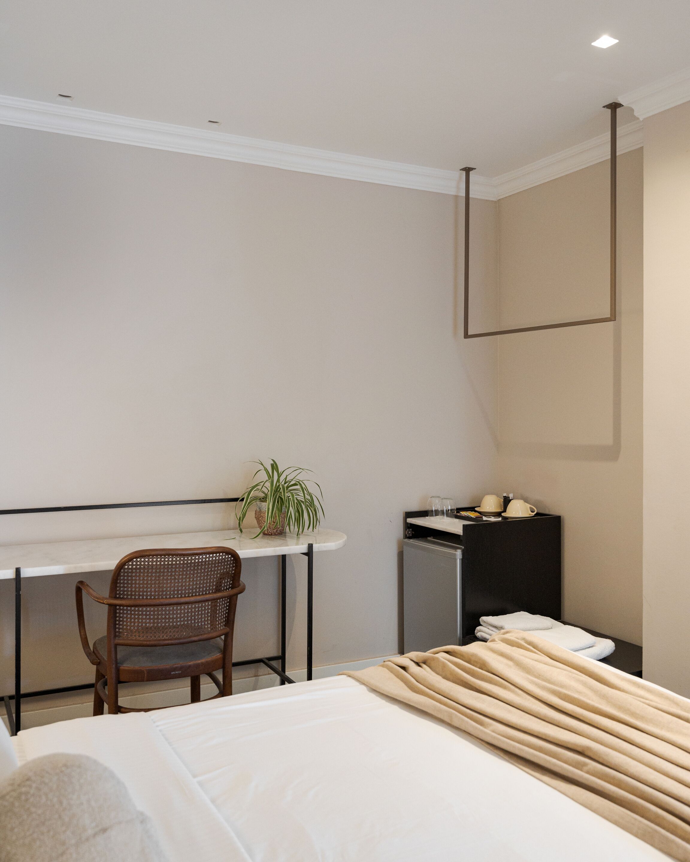 A modern hotel room with a bed, a desk, a chair, a small refrigerator, and a potted plant.