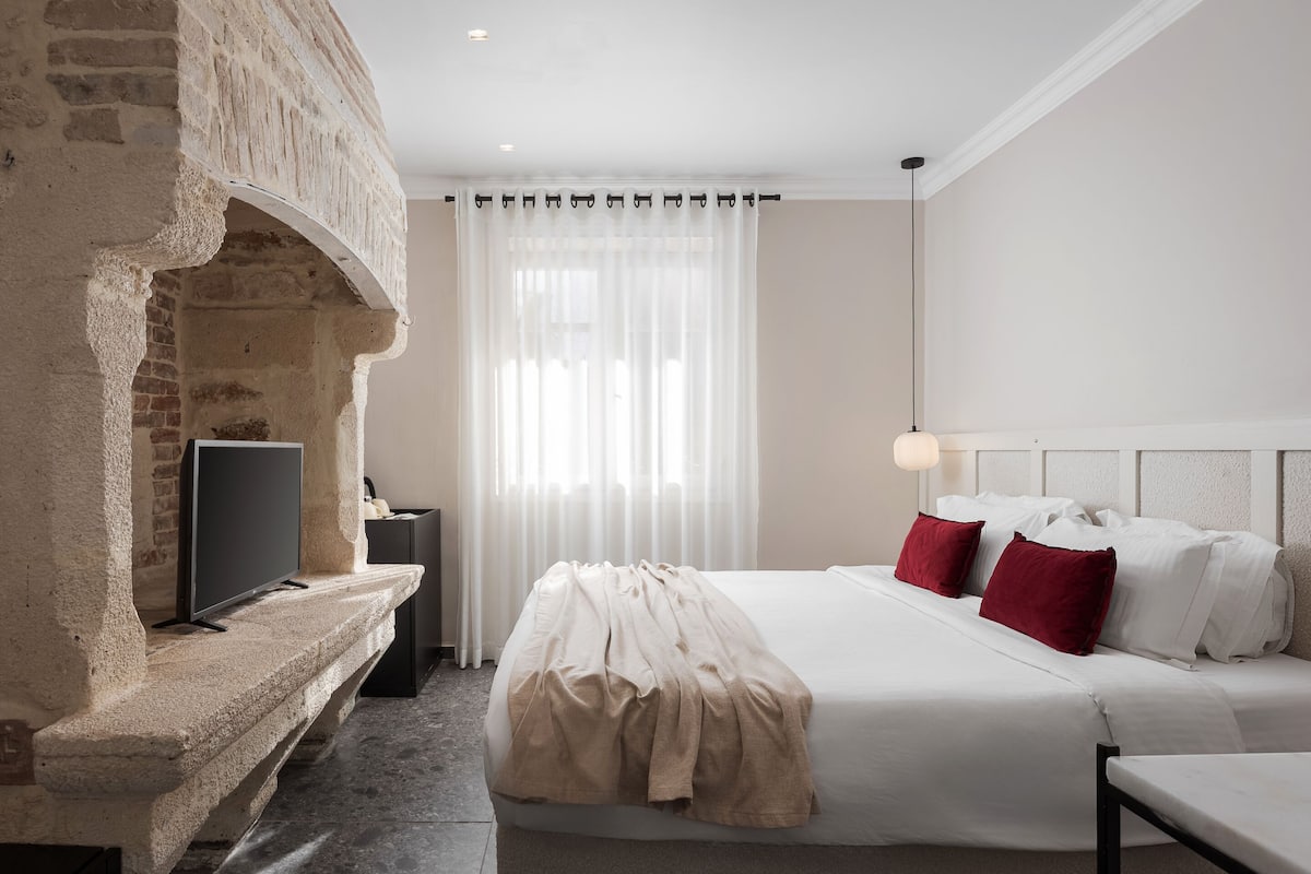 A hotel room with a stone fireplace, a bed with white and red pillows, a flat-screen TV on a stone shelf, and a large window with sheer curtains.
