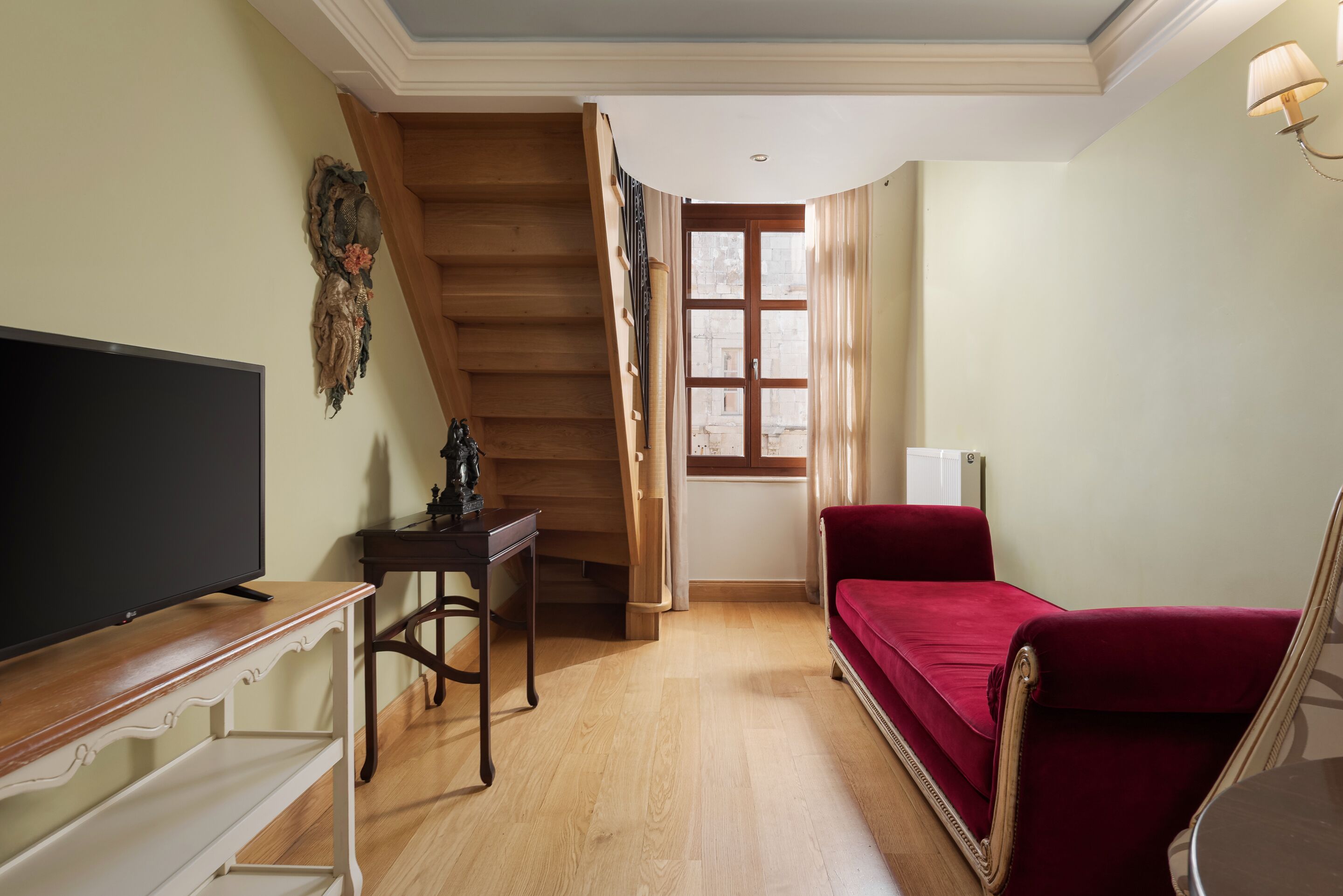 Una habitación con televisión sobre un mueble de madera, un escritorio, un sofá rojo y una escalera que lleva a otra habitación.