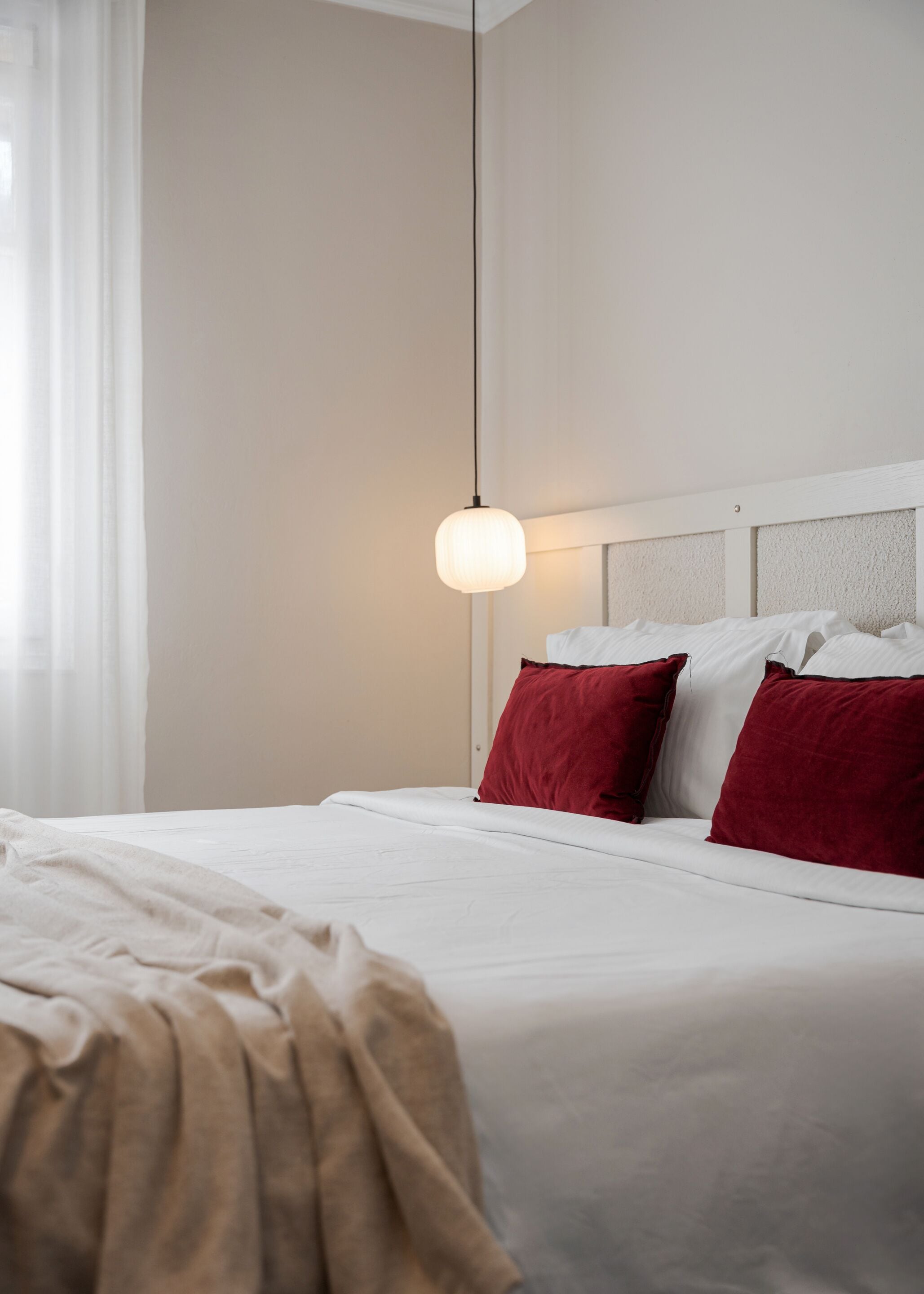 A hotel room with a bed, white bedding, red pillows, a hanging light fixture, and a window with sheer curtains.