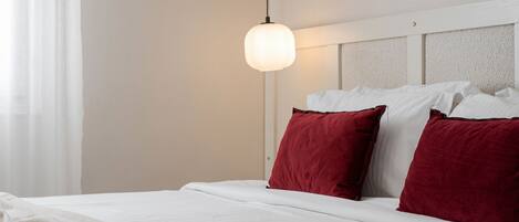 A hotel room with a bed, white bedding, red pillows, a hanging light fixture, and a window with sheer curtains.
