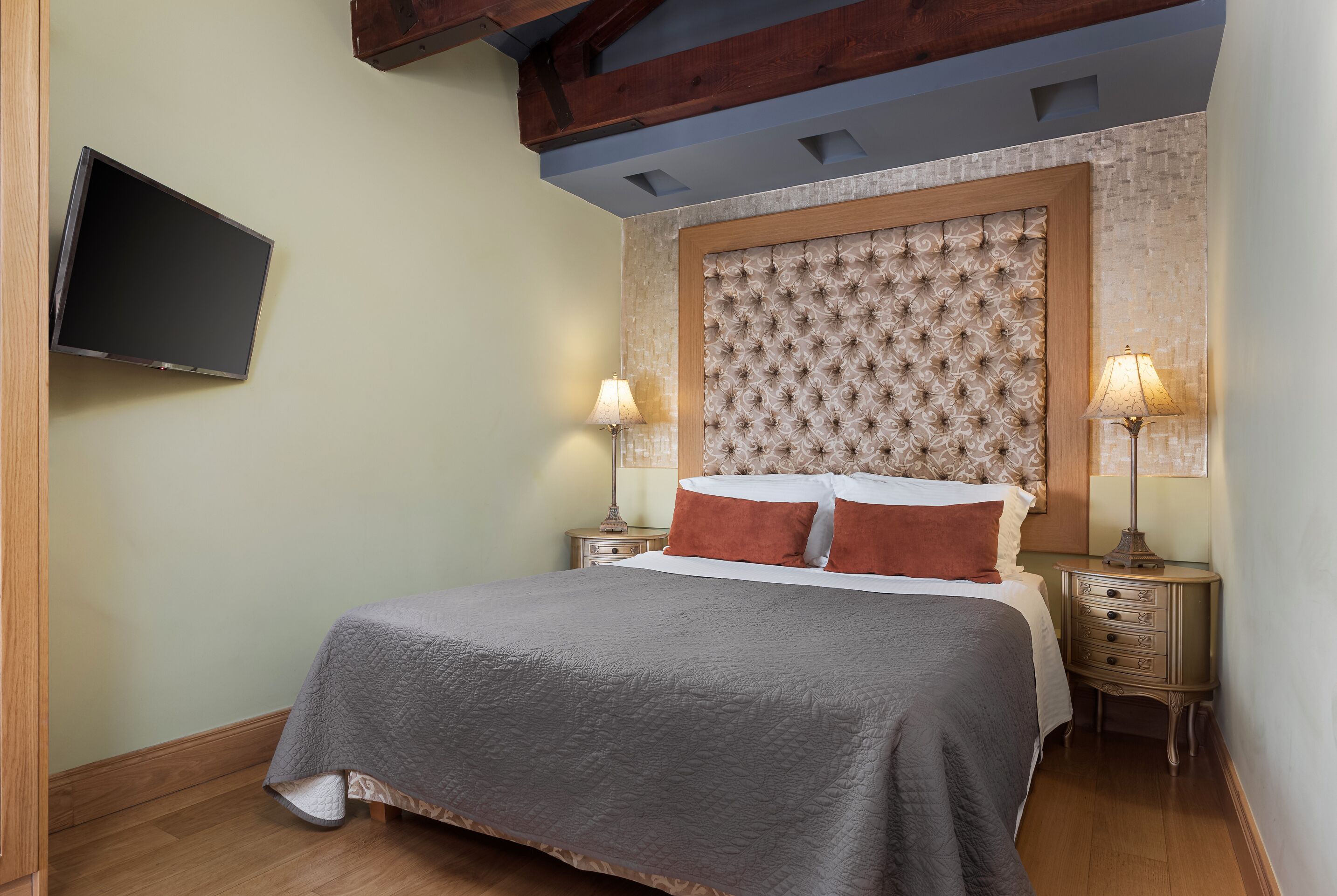 A bedroom with a bed, two bedside tables with lamps, a flat-screen TV mounted on the wall, and a decorative headboard.