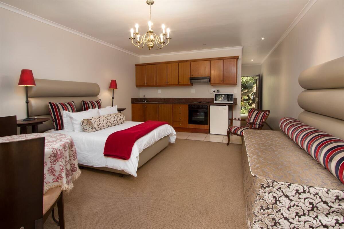 A hotel room with a bed, bedside tables, a sofa, a dining area with a kitchen, and a chandelier.