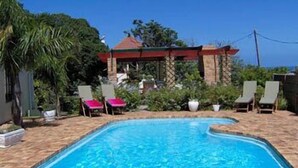 A rectangular swimming pool with a tiled deck, surrounded by lounge chairs and a covered area with a red roof. - Milkwood Lodge - Hermanus (Hermanus)