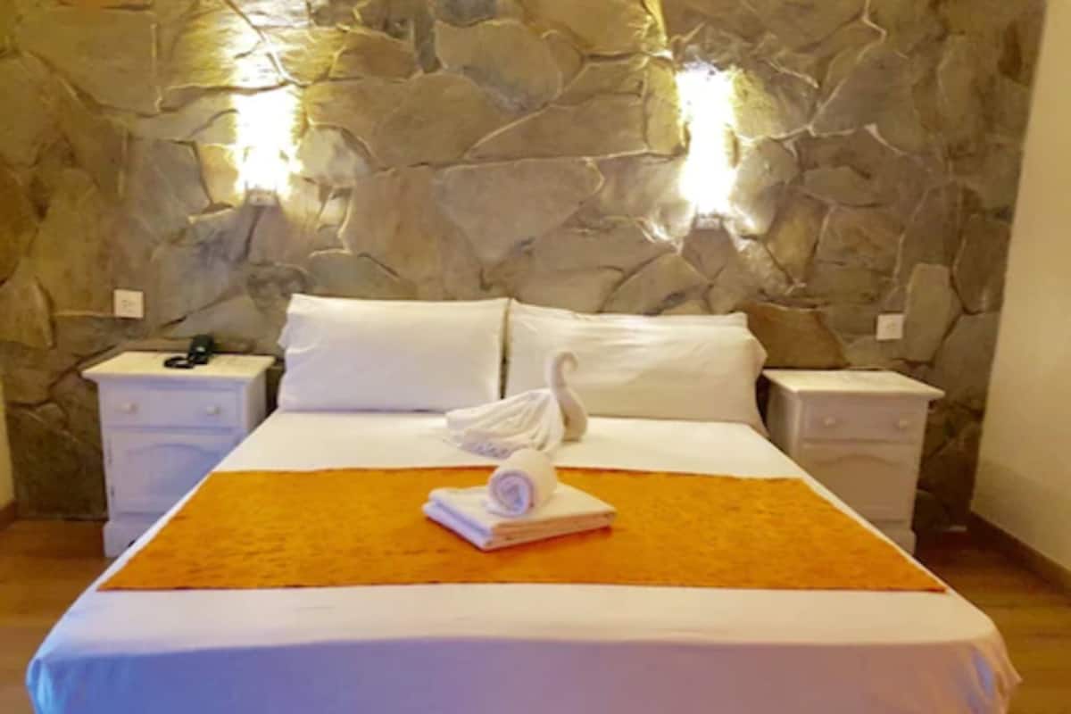 Habitación de hotel con pared de piedra, una cama con ropa blanca, una toalla doblada sobre la cama y dos mesitas de noche con lámparas.