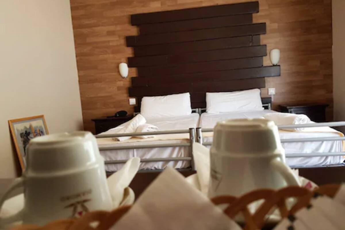Habitación de hotel con cabecera de madera, dos camas, una mesita de noche y una mesa con dos tazas blancas tipo termo.