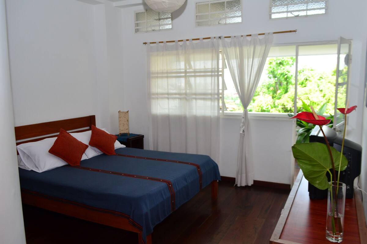 A bedroom with a wooden bed, white and blue bedding, a nightstand, a window with a view of greenery, and a potted plant.
