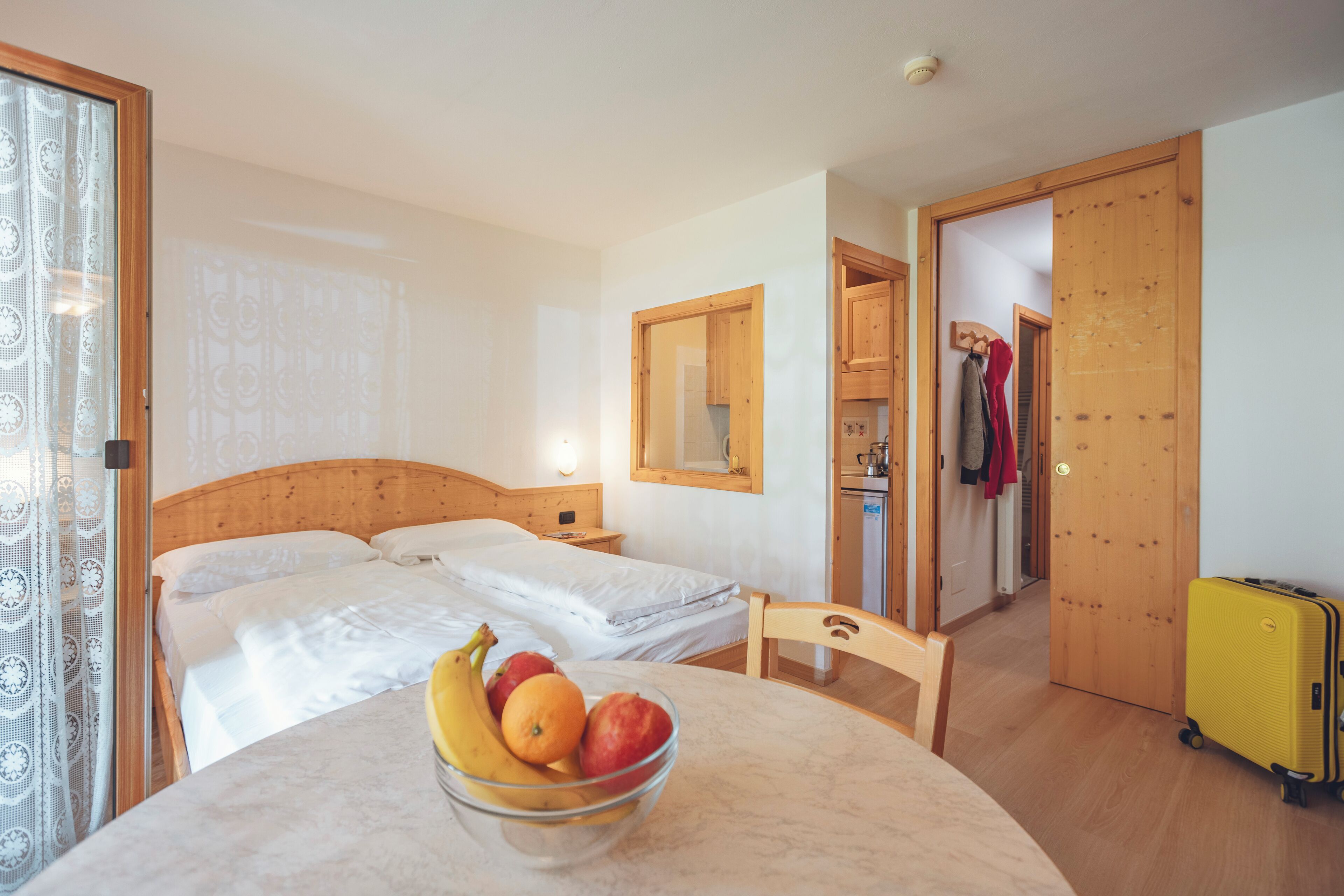 A hotel room with a bed, a table with a fruit bowl, a wooden chair, a yellow suitcase, and a kitchen area visible through an open door.