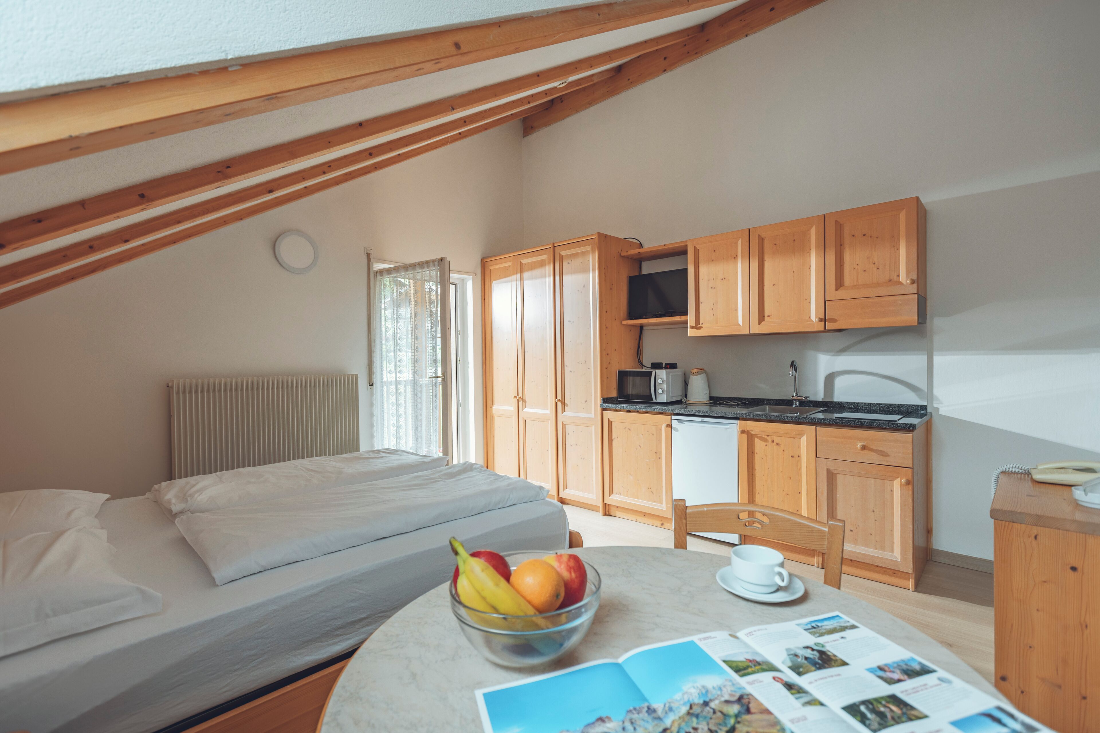 Une chambre avec un lit, une table de chevet sur laquelle se trouve un bol de fruits et un magazine. Il y a également une petite cuisine équipée avec des placards en bois et un four à micro-ondes.