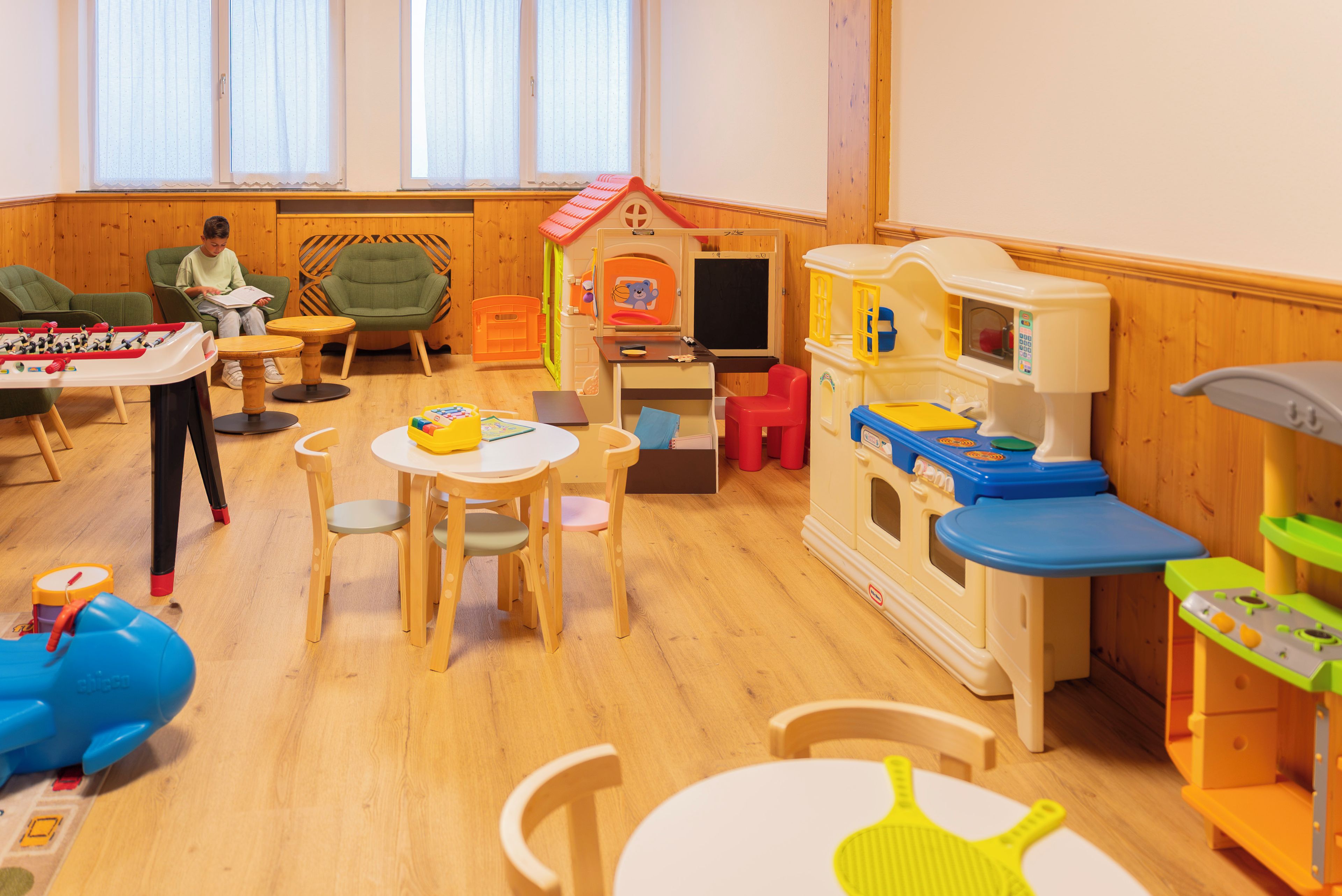 A children's playroom with wooden flooring, a table with chairs, and various play equipment including a toy kitchen set.