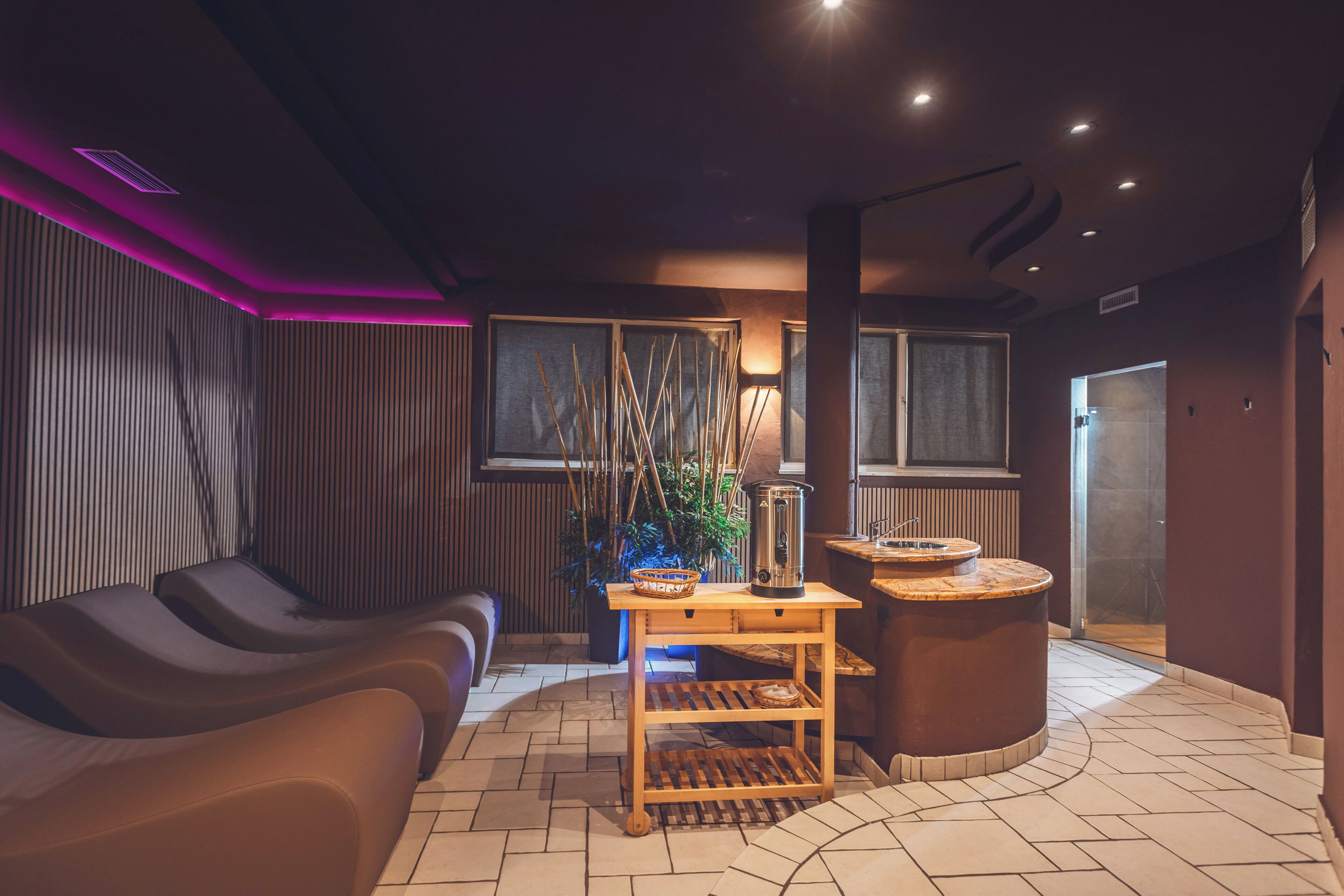 A modern spa room with a massage chair, a wooden table with a basket, a water feature, and a stone basin.
