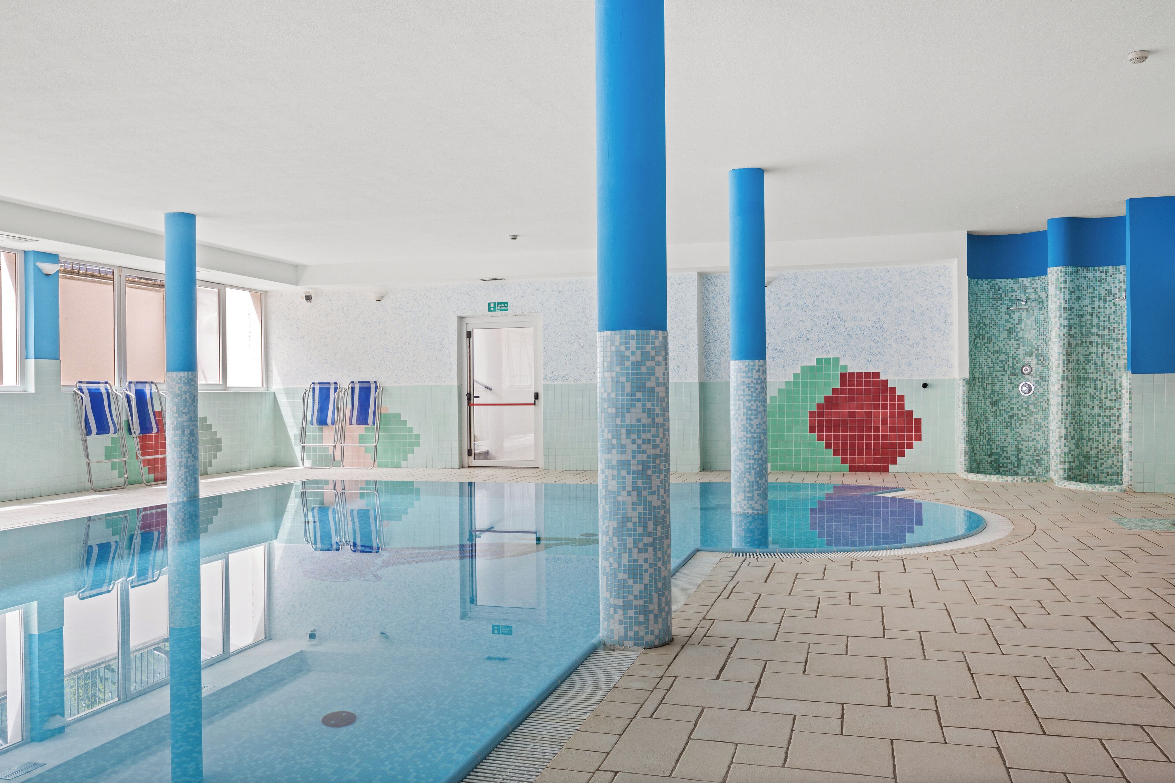 An indoor swimming pool with blue-tiled walls and columns, clear water, and a tiled floor.