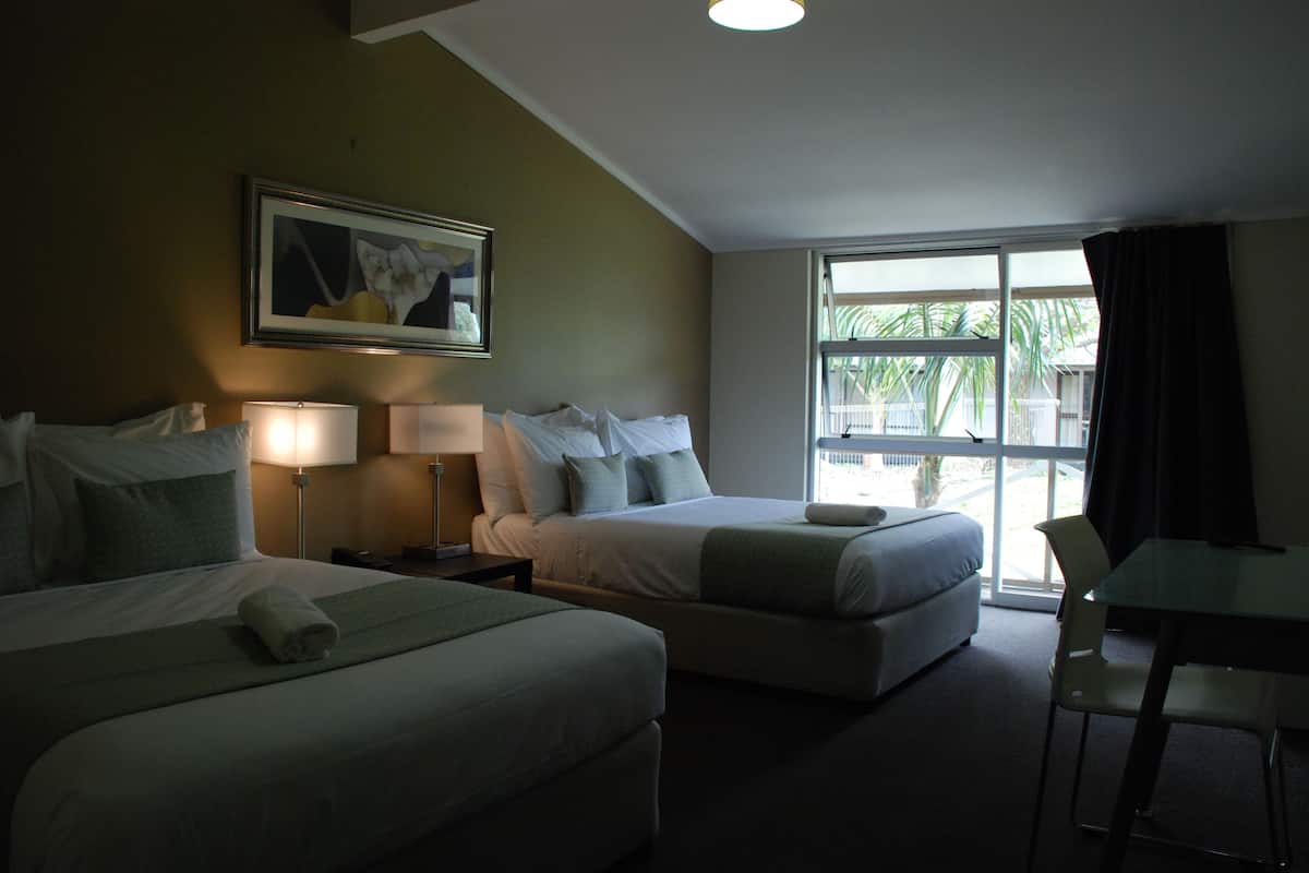 A hotel room with two beds, a desk, and a window with a view of palm trees.