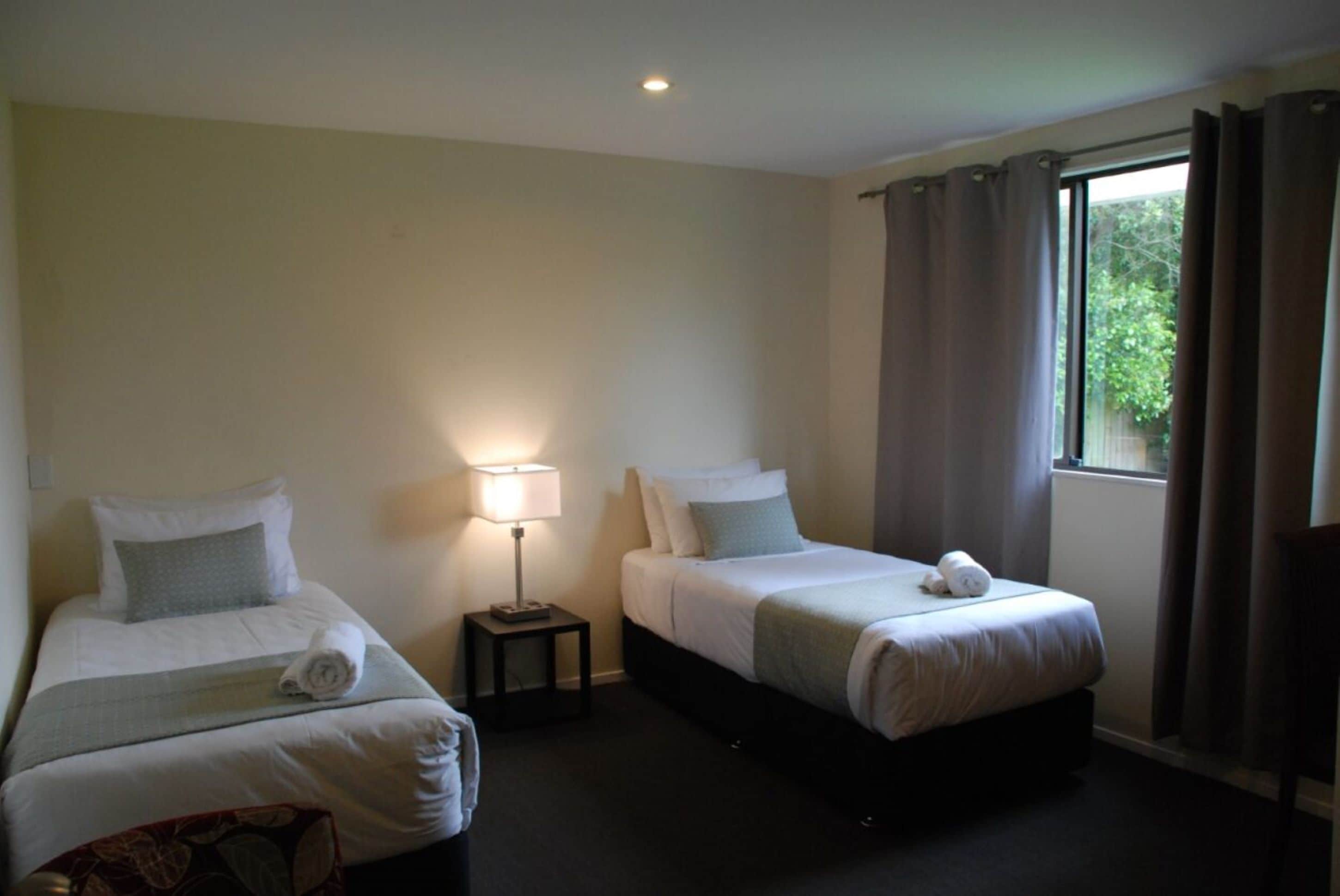 Habitación de hotel con dos camas individuales, una mesita de noche con lámpara, una ventana con cortinas y vistas al exterior con vegetación.