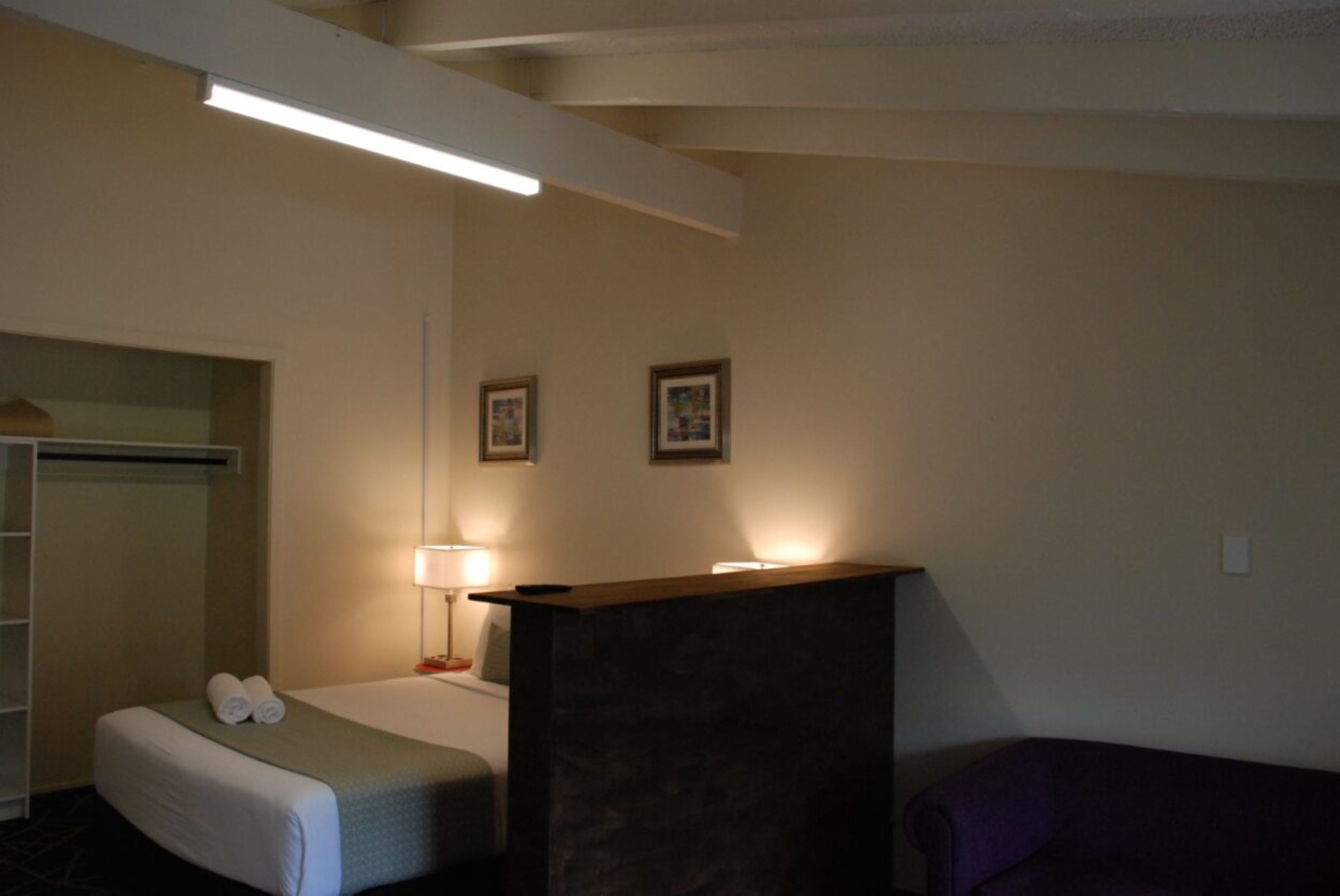 Una habitación de hotel con cama, escritorio, silla, armario y dos cuadros en la pared.