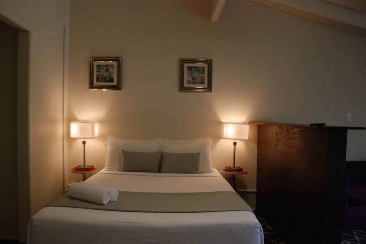 A hotel room with a bed, two bedside lamps, and two framed pictures on the wall.