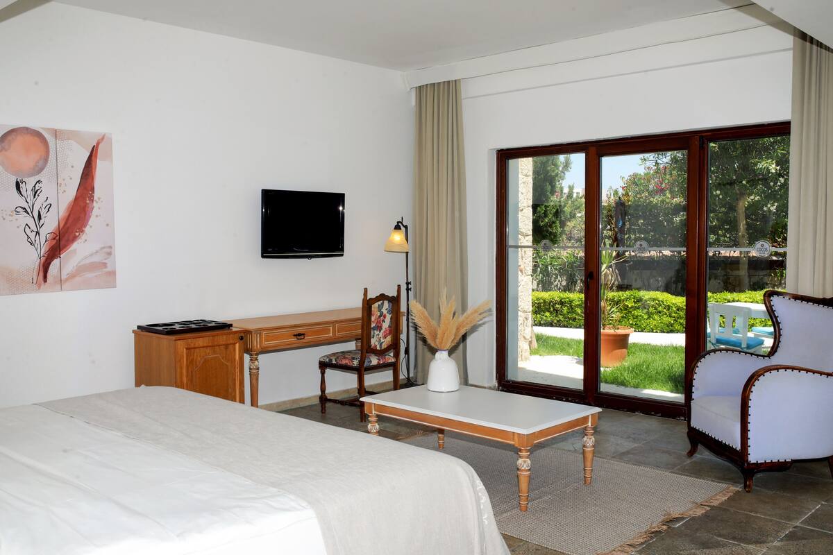 Une chambre d’hôtel équipée d’un lit, d’un bureau, d’une chaise, d’une télévision et offrant une vue sur le jardin grâce à la porte vitrée.