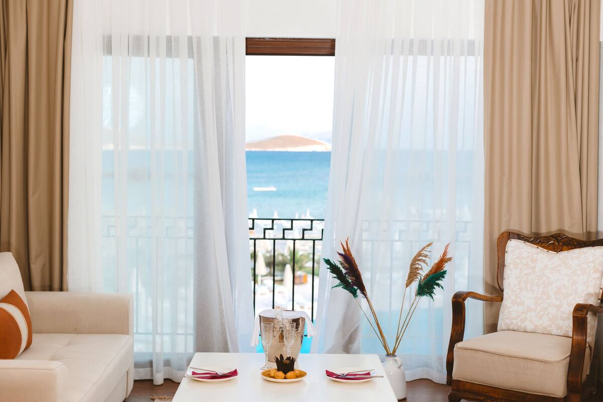 Une pièce avec vue sur la mer, une table blanche ornée d’un vase et d’une assiette de fruits, et une chaise en bois.