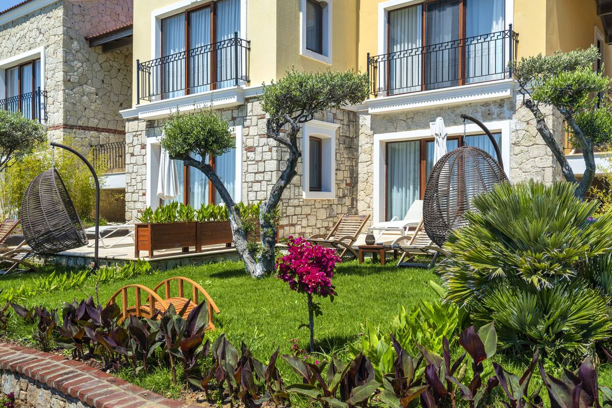 Un hôtel à la façade en pierre, doté de balcons et d’un jardin bien entretenu, agrémenté de diverses plantes et d’espaces aménagés pour se détendre.