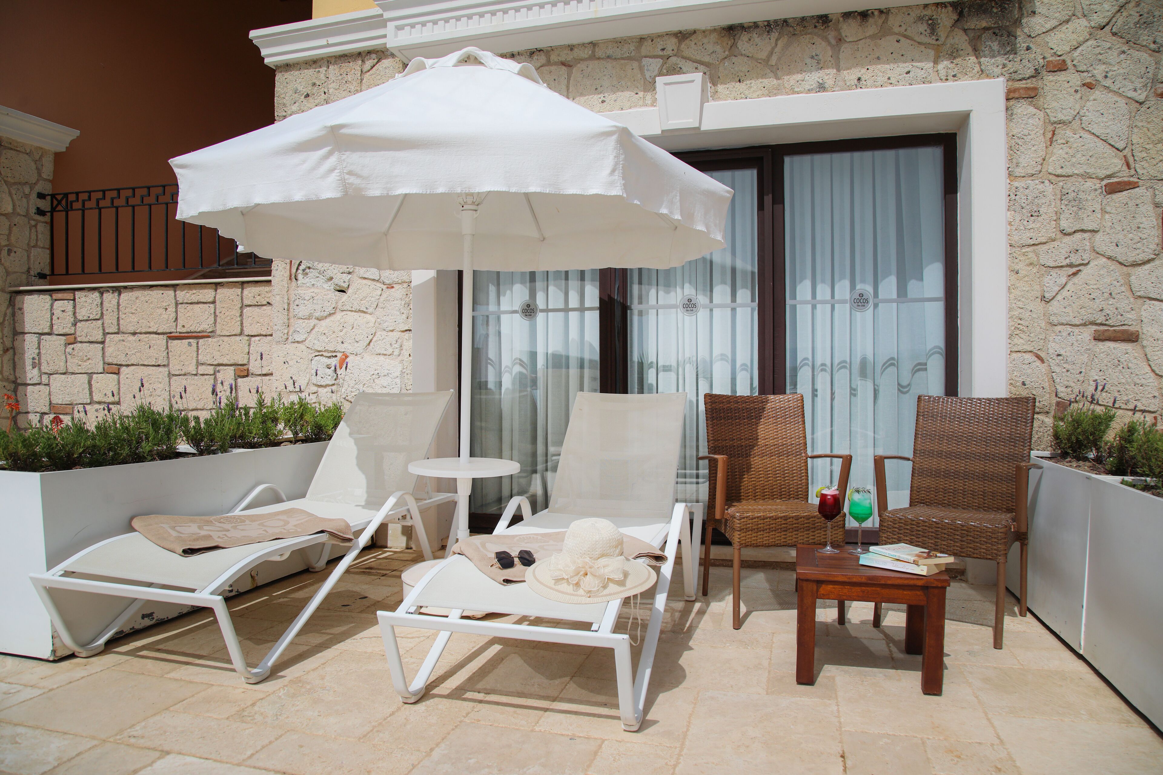A patio with white lounge chairs, a table with a drink, and an umbrella.