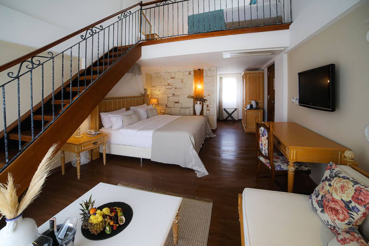 Une chambre avec un grand lit, un escalier en bois, un téléviseur fixé au mur et un coin repas avec une table et des chaises.