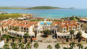 Um resort com piscina central, várias villas com telhados vermelhos e vista para o mar e turbinas eólicas ao fundo.