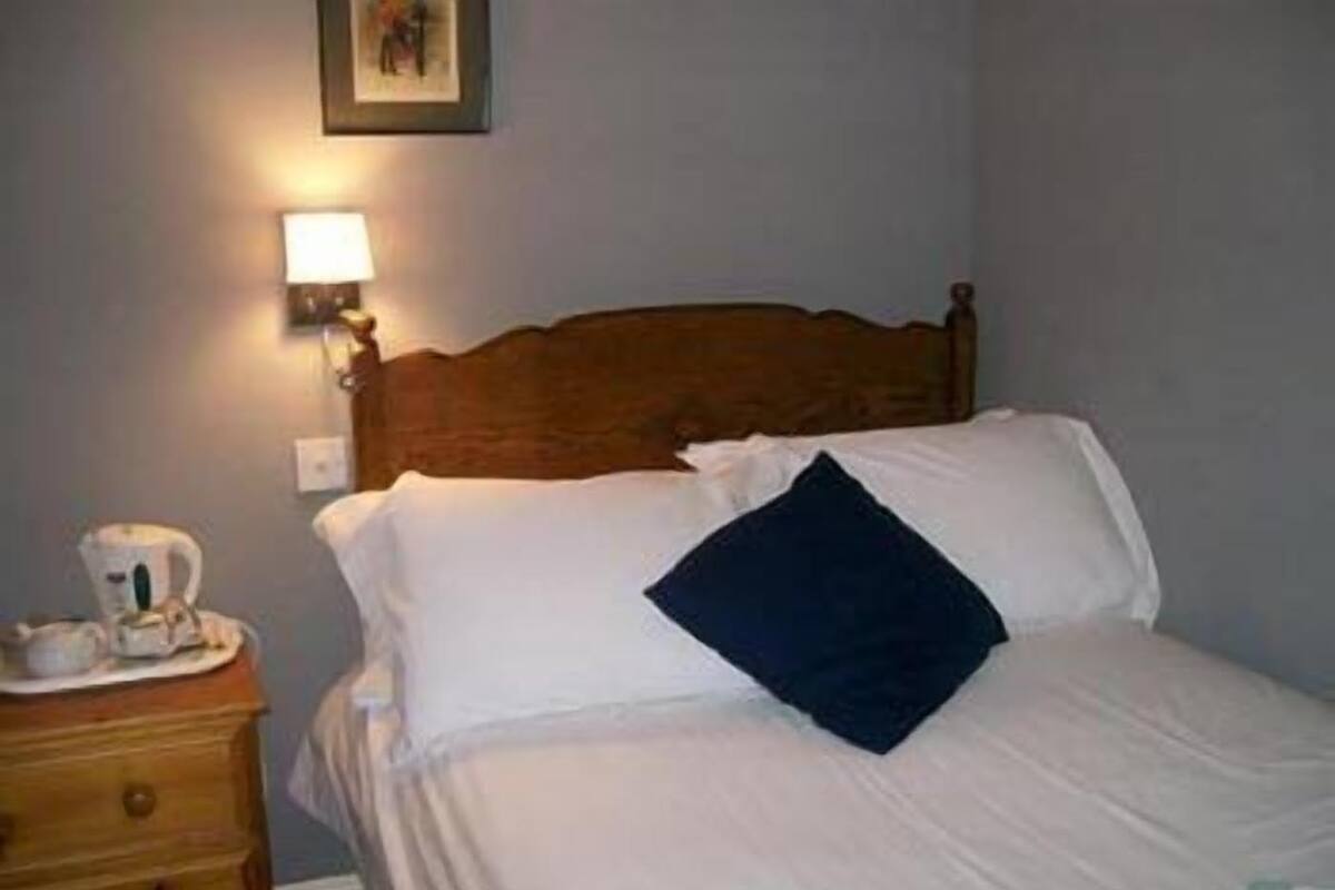 A neatly made bed with white linens and a dark blue pillow, a bedside table with a cup and saucer, and a wall-mounted lamp.