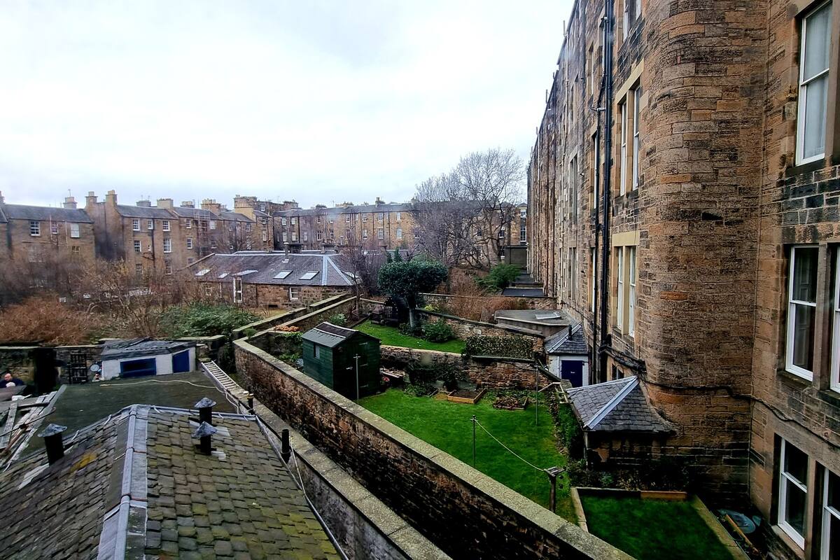 A view of a courtyard with a stone wall, a small garden, and a building with multiple windows.