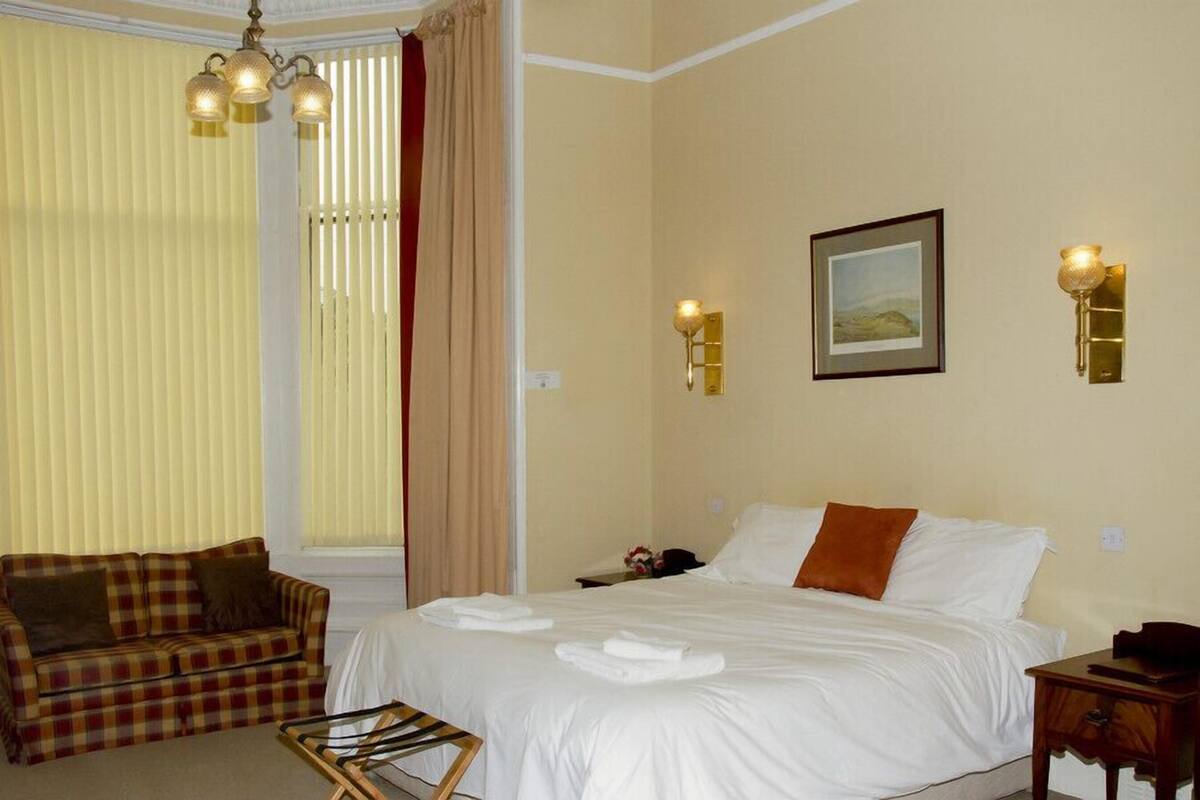 A hotel room with a bed, a sofa, a table with towels, and wall-mounted lamps.