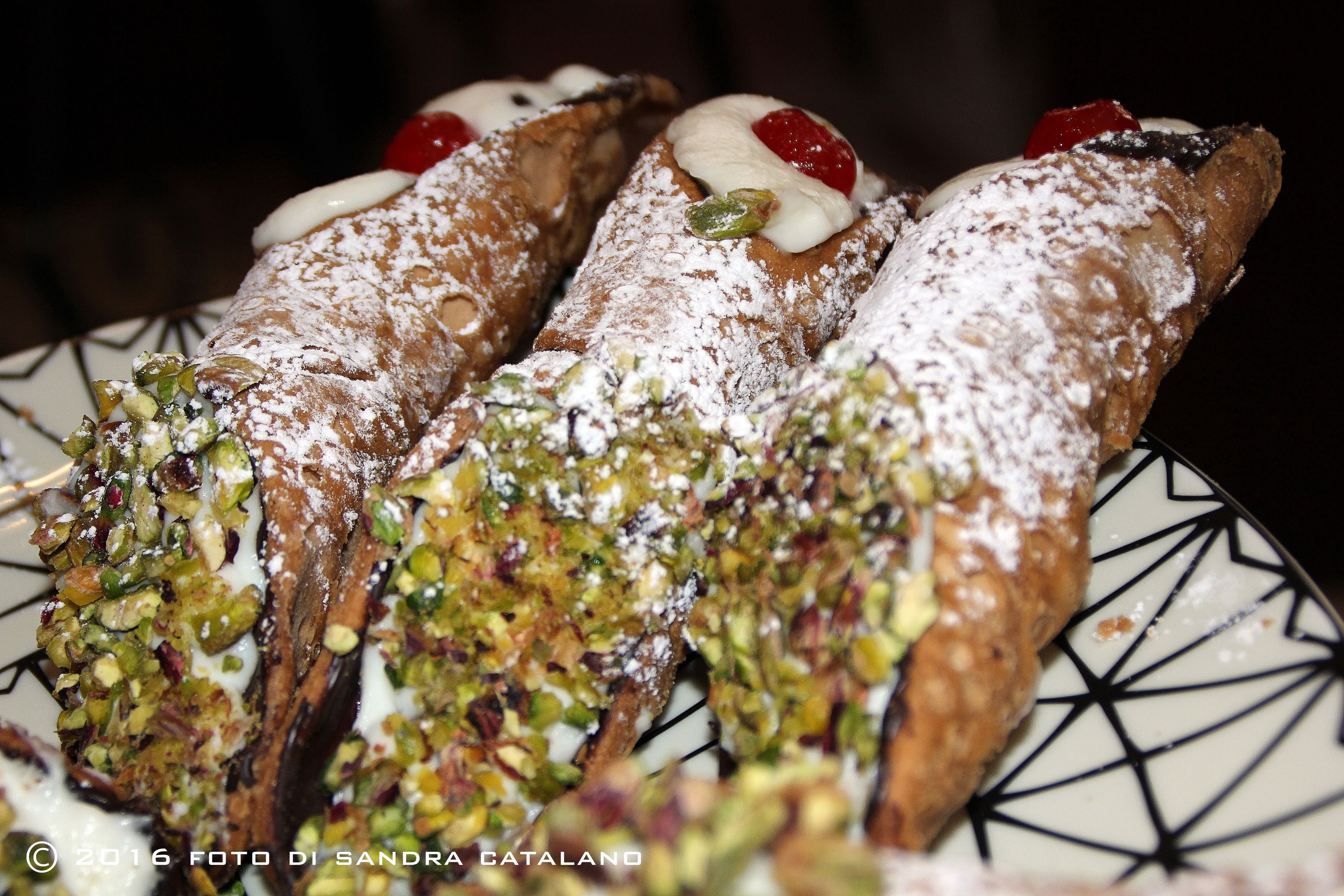Trois cannolis saupoudrés de sucre glace et garnis de pistaches, présentés sur une assiette à motifs.