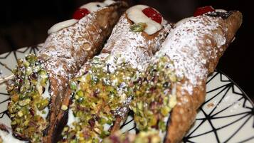 Trois cannolis saupoudrés de sucre glace et garnis de pistaches, présentés sur une assiette à motifs.