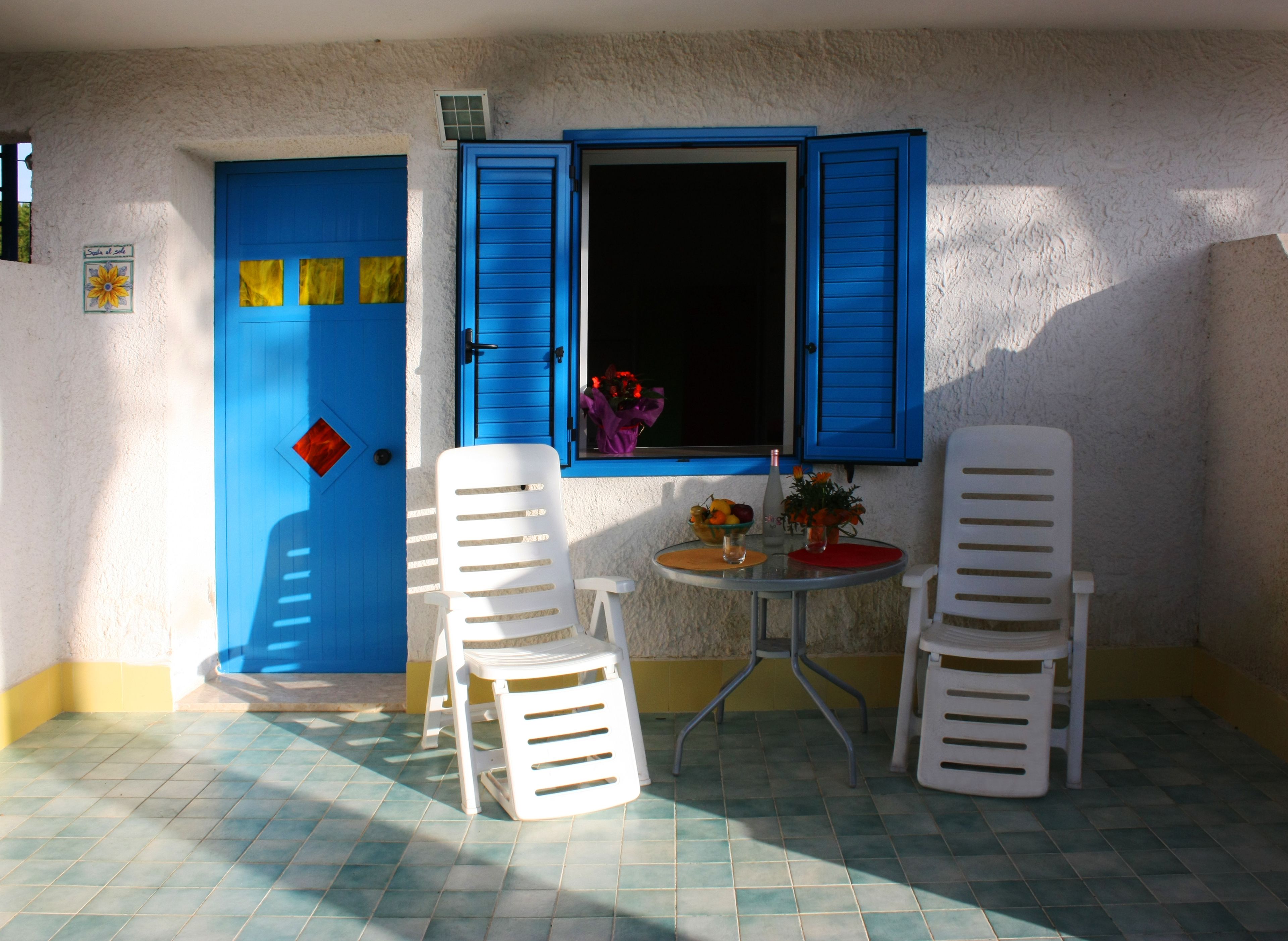 Een klein buitenzitje met twee witte plastic stoelen en een ronde tafel, een blauwe deur en een raam met blauwe luiken.