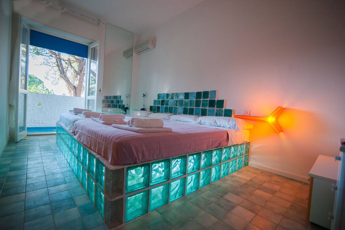 Ein Schlafzimmer mit einem Bett, einem Nachttisch, einem Spiegel und einem Fenster.