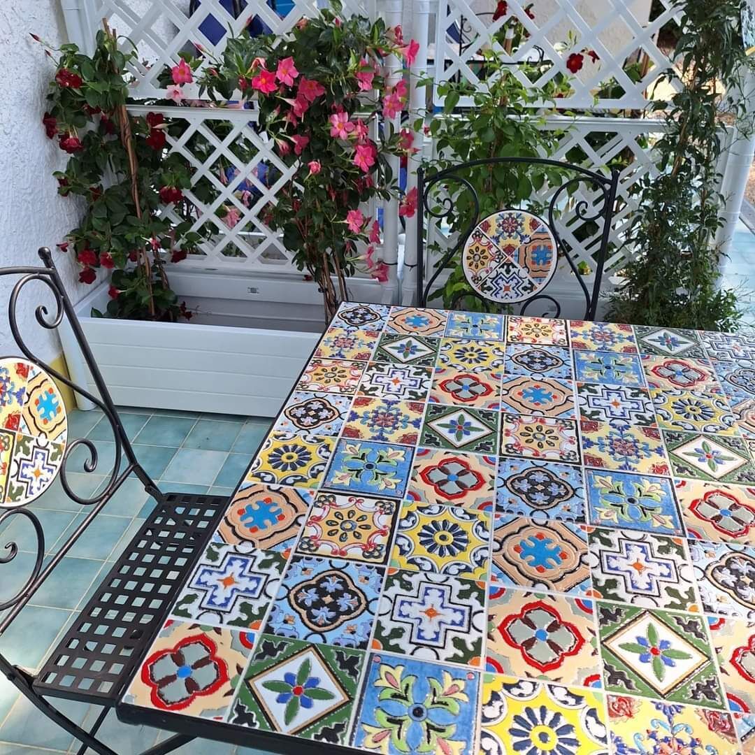 A table with a colorful tiled top, surrounded by a white lattice fence and potted plants.