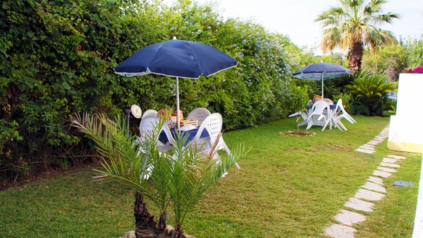 An outdoor seating area with tables and chairs under umbrellas, surrounded by lush greenery and a palm tree. - Suite House L'isola che non c'è (Syracuse)