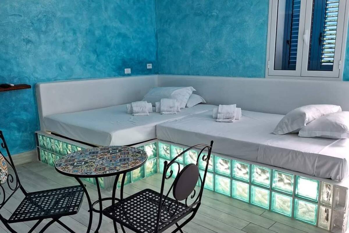 Ein Zimmer mit blauer Wand, einem weißen Sofa, zwei Metallstühlen, einem kleinen runden Tisch und einem Fenster mit blauen Fensterläden.